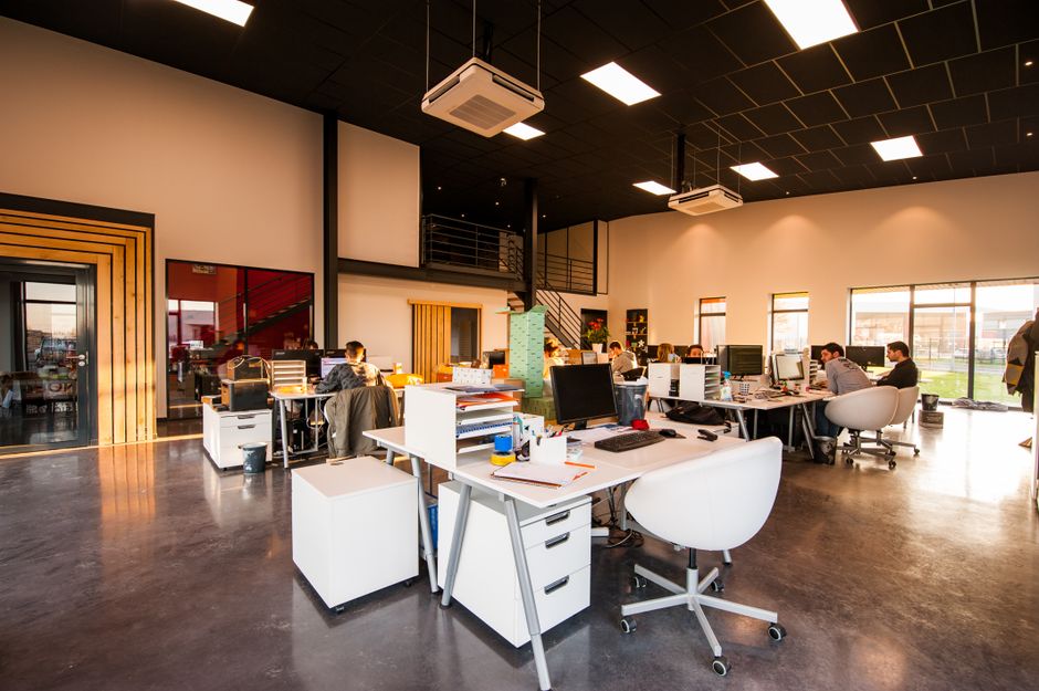Open-plan office with white desks, computers, and employees at workstations under bright ceiling lights.